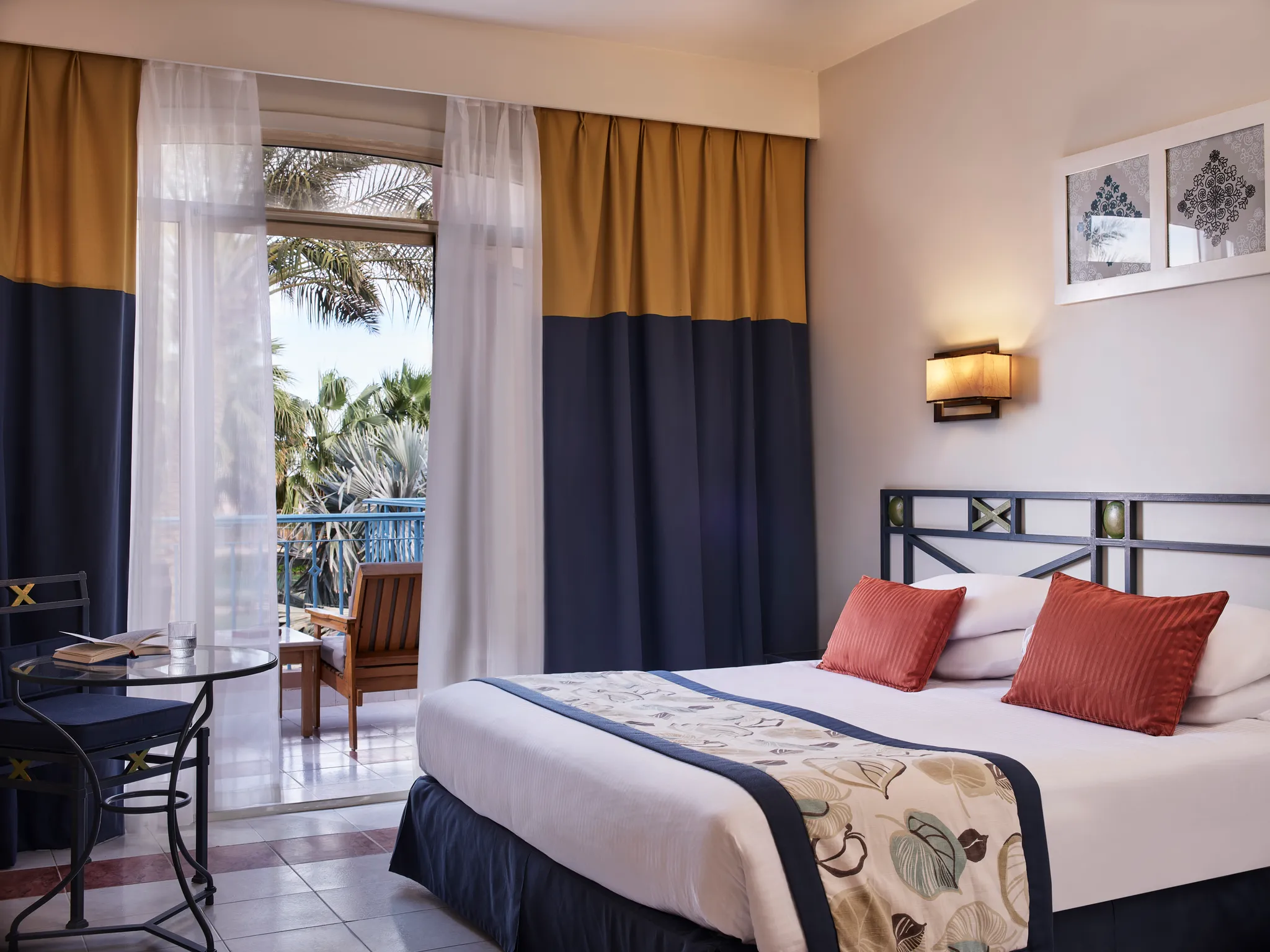 Detailansicht des Kingsize-Bettes in der Paradise Suite mit gemustertem Bettläufer, roten Akzentkissen und einem angrenzenden Glastisch am Terrassenfenster im Club Paradisio Hotel El Gouna