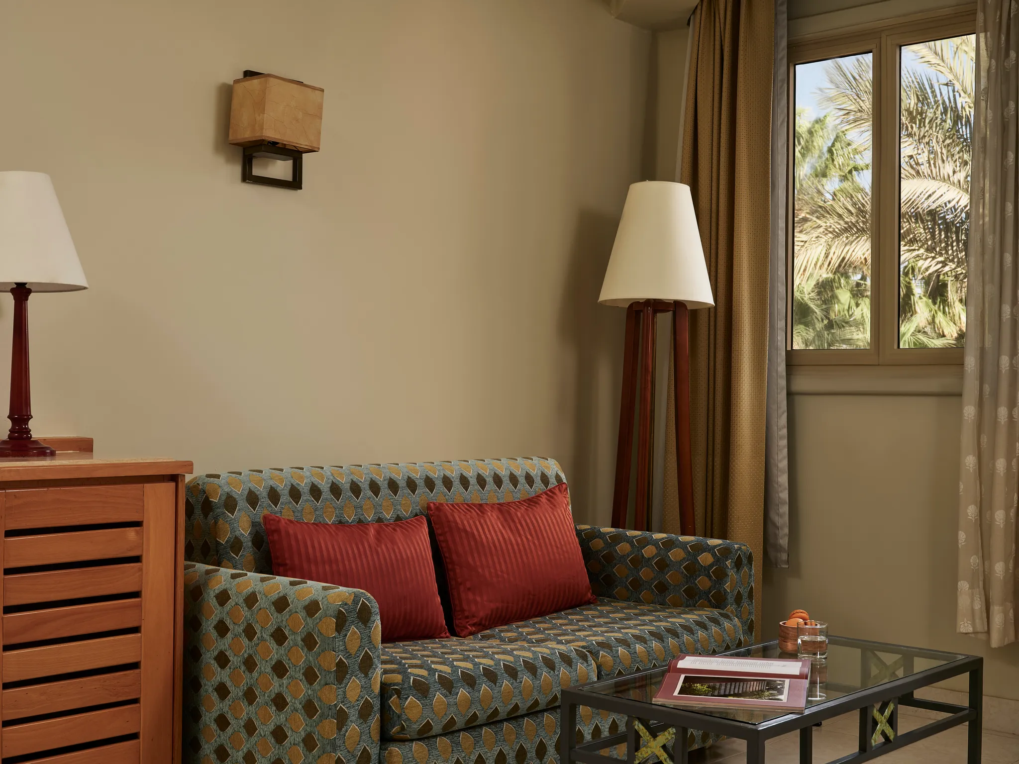 Wohnbereich in der Paradise Suite mit einem Sofa aus gemustertem Stoff, roten Kissen, einer Standlampe auf Dreifuß und einem Couchtisch am Fenster im Club Paradisio Hotel El Gouna