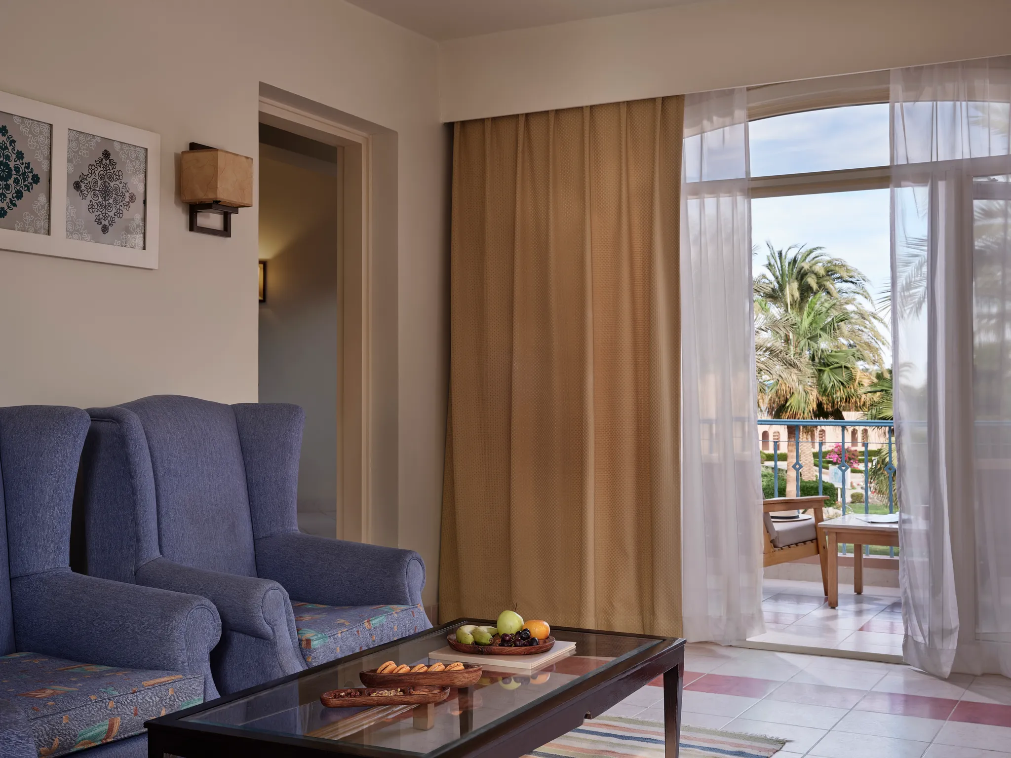 Wohnzimmer in der Paradise Suite mit blauen Sesseln, einem gläsernen Couchtisch mit Obstplatte und einer Balkontür mit Blick auf die Resortgärten des Club Paradisio Hotels in El Gouna