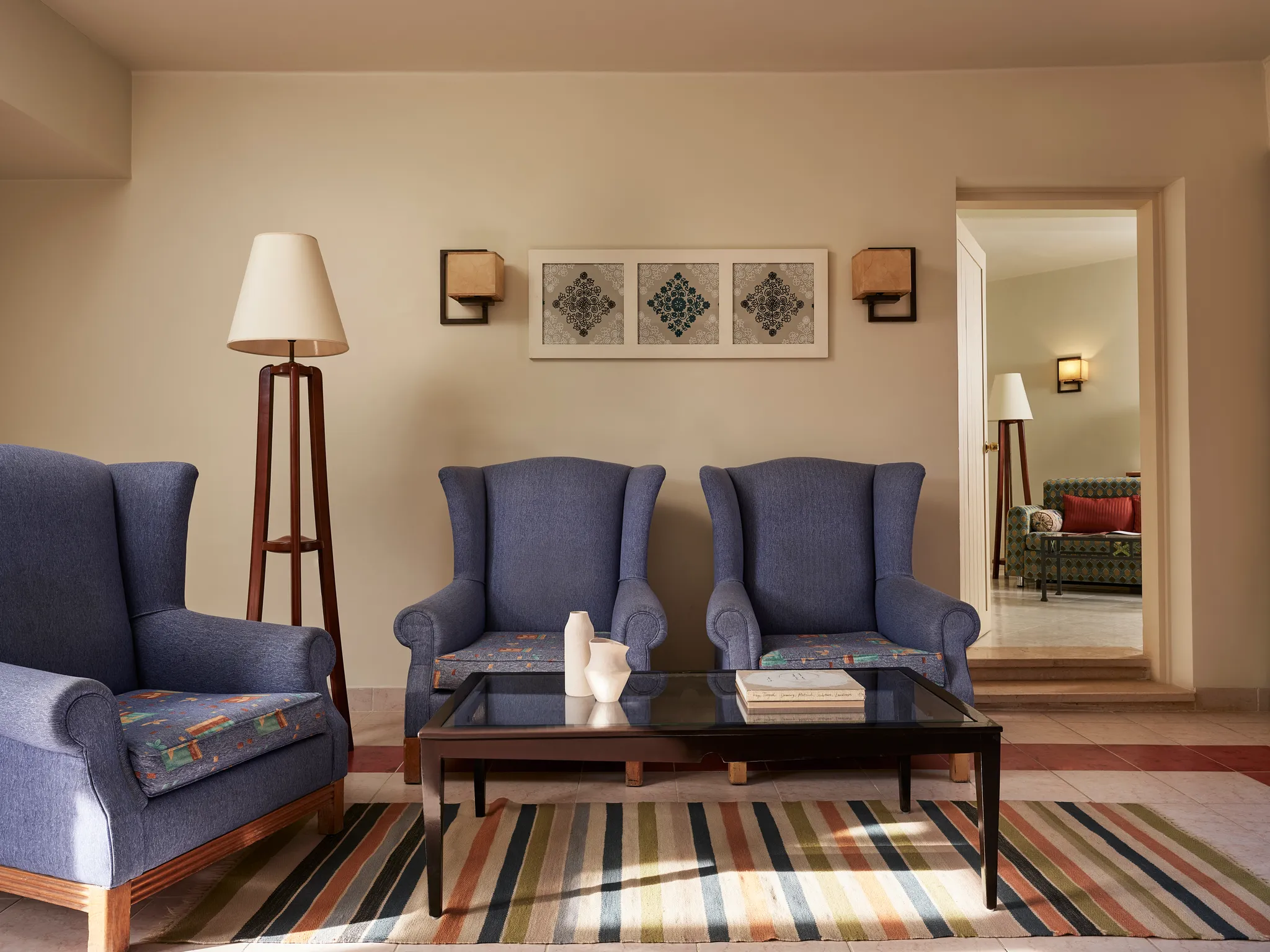 Komplette Wohnzimmerausstattung in der Paradise Suite mit drei blauen Sesseln, einer Stativ-Stehlampe, einem gläsernen Couchtisch auf einem gestreiften Teppich sowie einer Verbindungstür zum angrenzenden Zimmer im Club Paradisio Hotel El Gouna