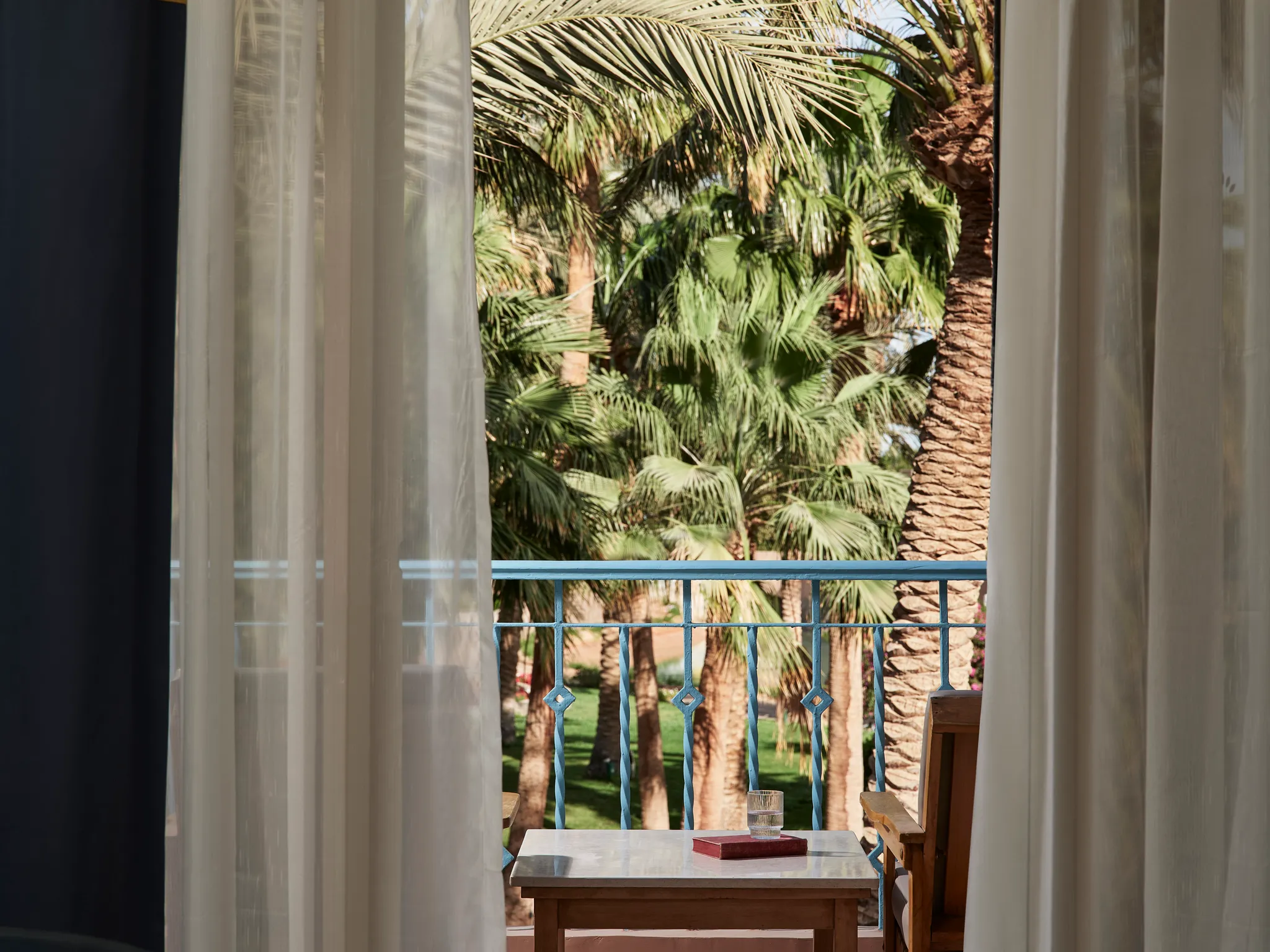 Nahaufnahme des Balkons der Paradise Suite durch hauchdünne weiße Vorhänge mit Blick auf die blauen Geländer und die üppig grünen Palmen im Club Paradisio Hotel El Gouna