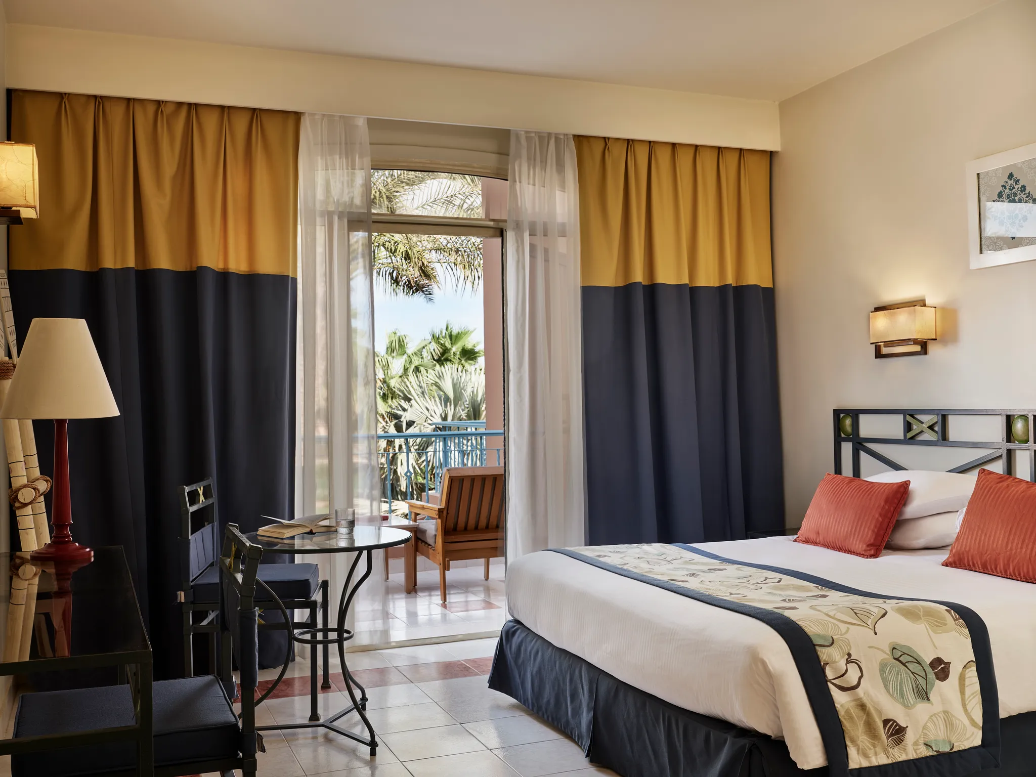 Helles Schlafzimmer in der Paradise Suite mit einem luxuriösen Kingsize-Bett, zweifarbigen Vorhängen, einer kleinen Esszimmergarnitur aus Glas und einer offenen Terrassentür mit Blick auf die Resortgärten des Club Paradisio Hotels in El Gouna