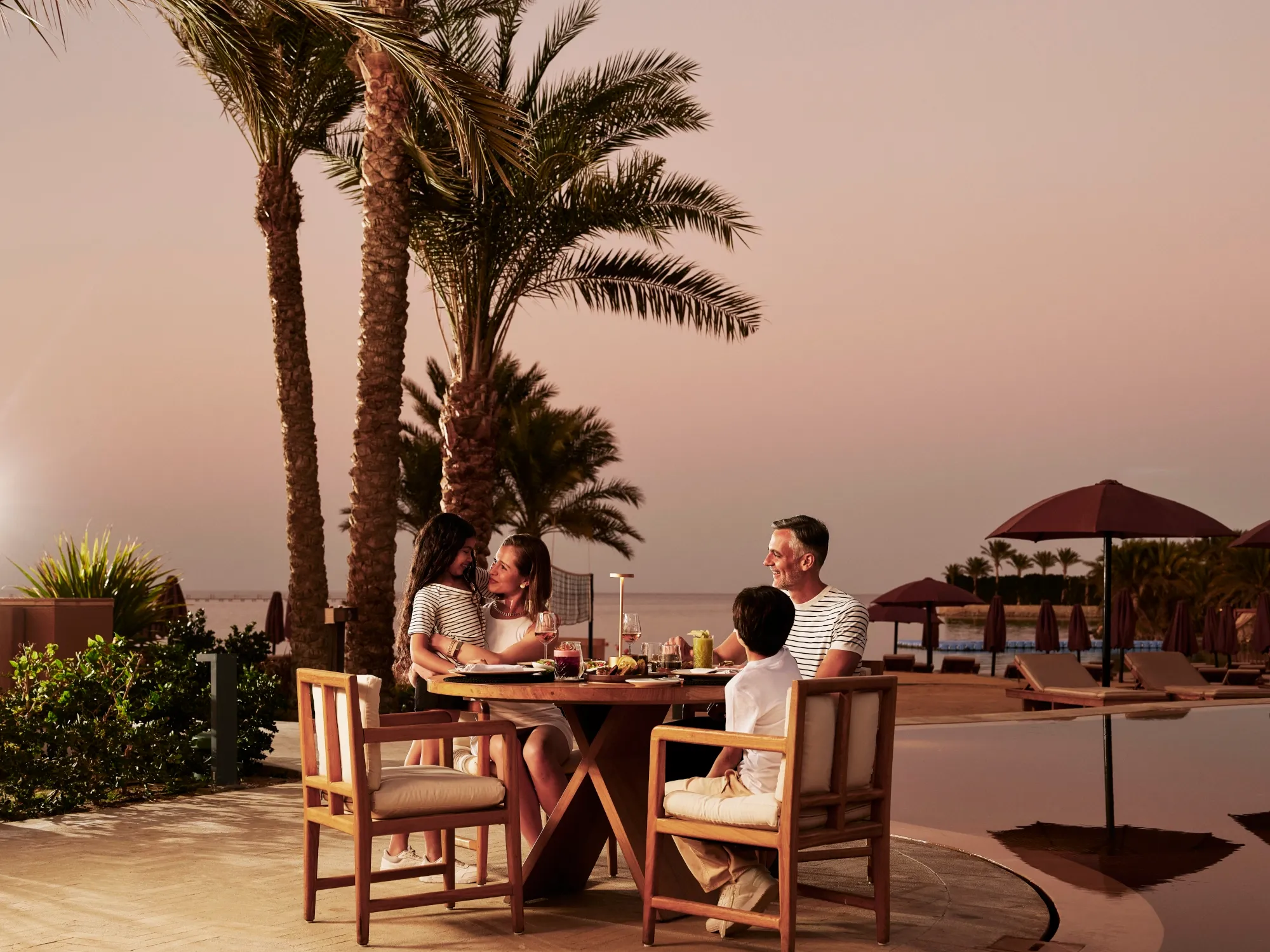 Family of four having dinner outdoors among palm trees at dusk, with a pool and beach cabanas in the background