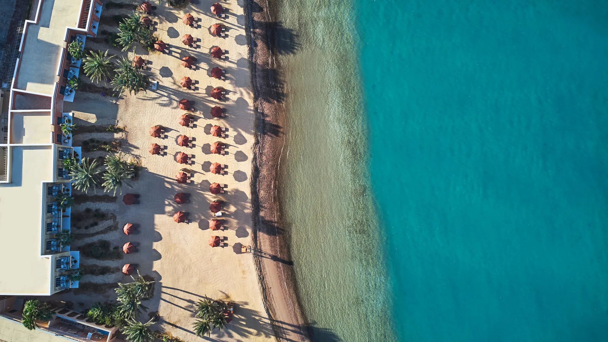 Aerial top-down view of the sandy beach lined with brown parasols, palm trees, and resort buildings alongside clear turquoise sea waters at The Chedi El Gouna